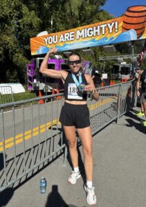 Runner wearing black shorts and sunglasses stands at the finish line, flexing a bicep and holding a phone, with a medal around their neck. Banner reads "YOU ARE MIGHTY!" in background.